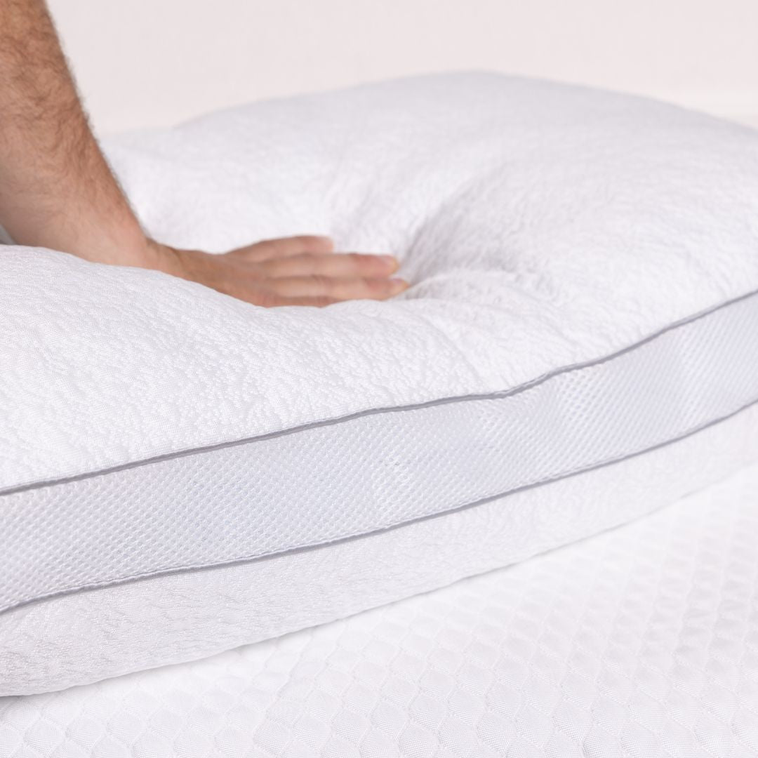 Close-up of a person's hand touching a white mattress topper with gray stitching on a light background.
