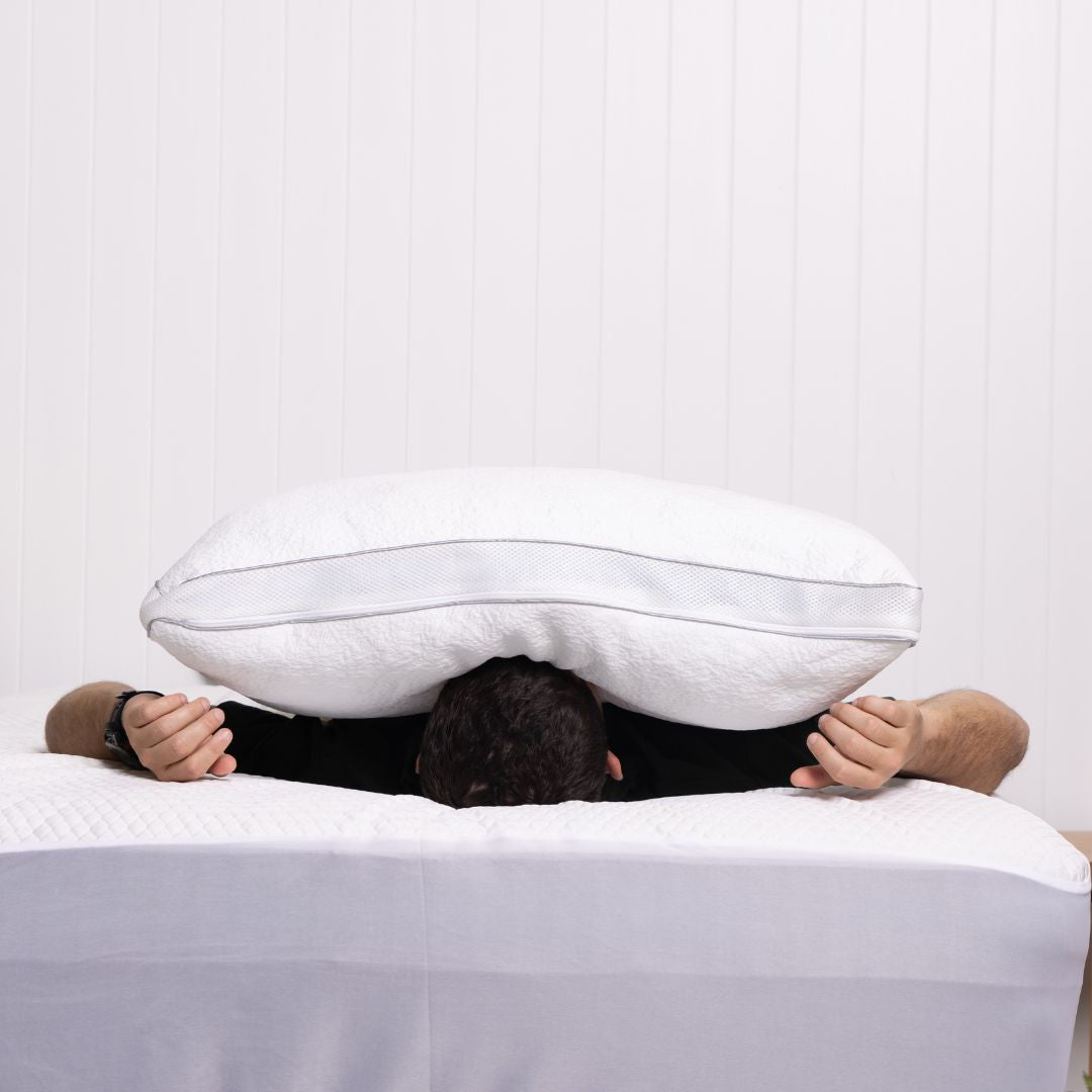 Person lying on a bed with a large white pillow over their head against a plain background
