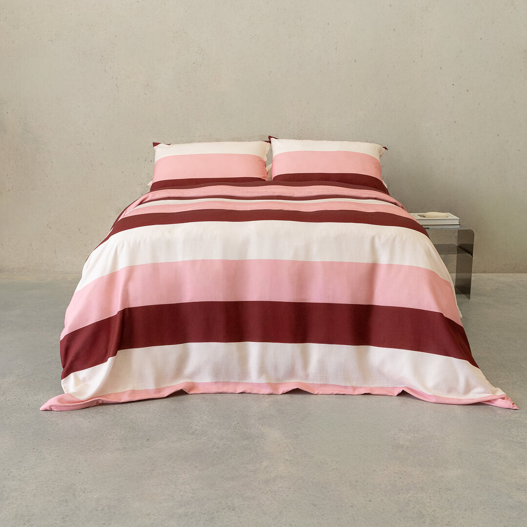 Pink, blush, cream, and burgundy striped quilt cover set with matching pillowcases displayed on a neatly made bed in a modern minimalist bedroom.