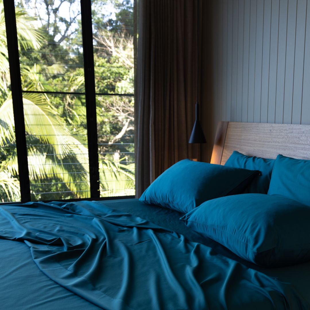 Bed with teal bedding in a room with large windows showing greenery outside.