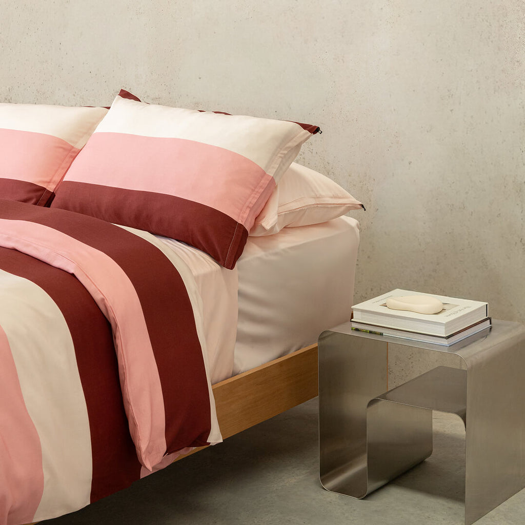 Close-up of a striped quilt cover in blush pink, cream, and burgundy tones paired with matching pillowcases and soft neutral sheets beside a contemporary metal bedside table.