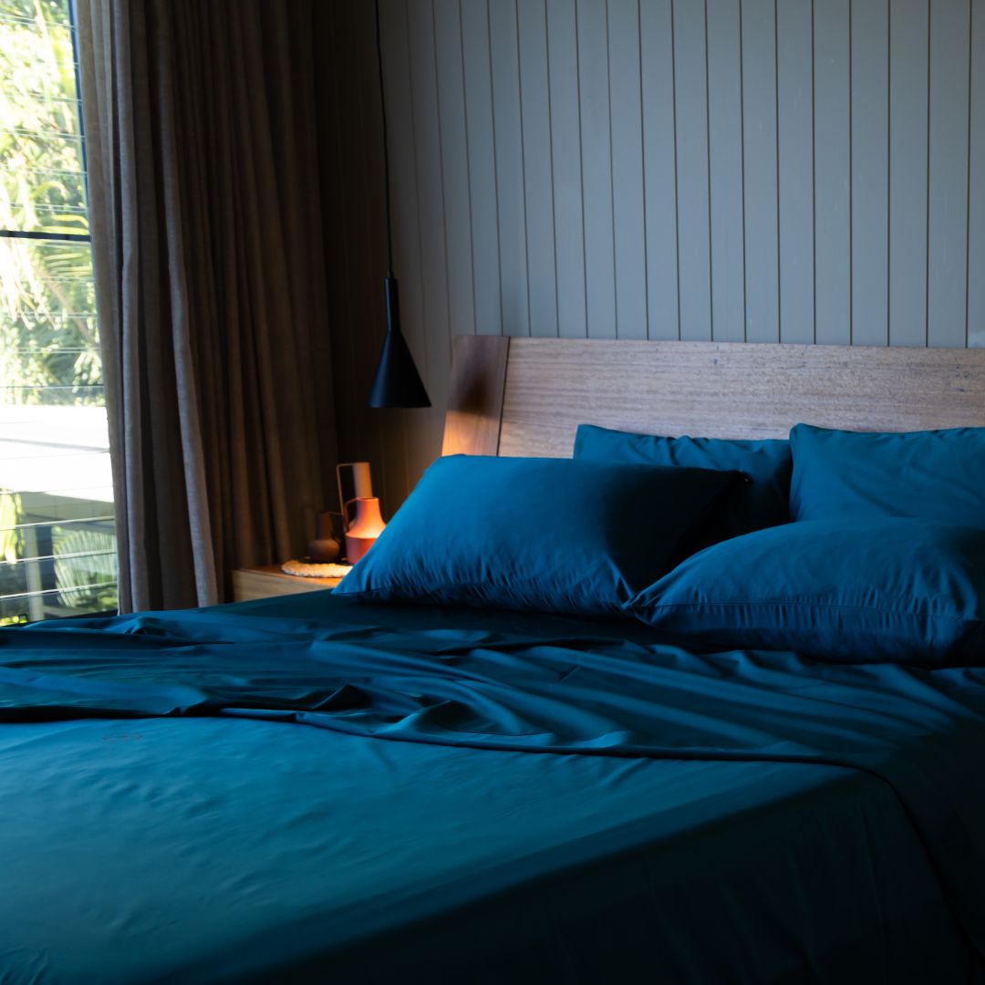 Bed with teal bedding in a room with large windows showing greenery outside.