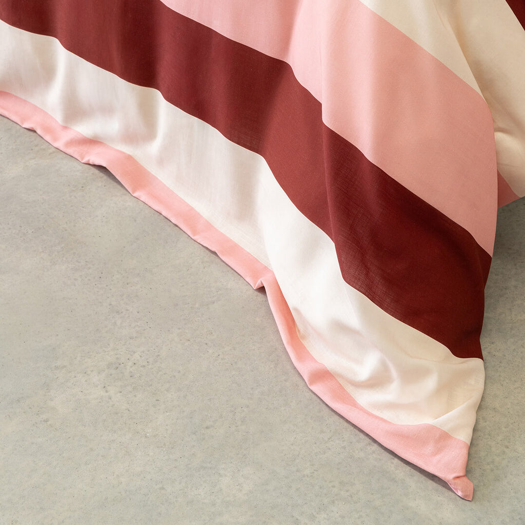 Close-up of a pink, cream, and burgundy striped quilt cover draped over the side of a bed, highlighting the soft fabric texture and bold horizontal stripe design.