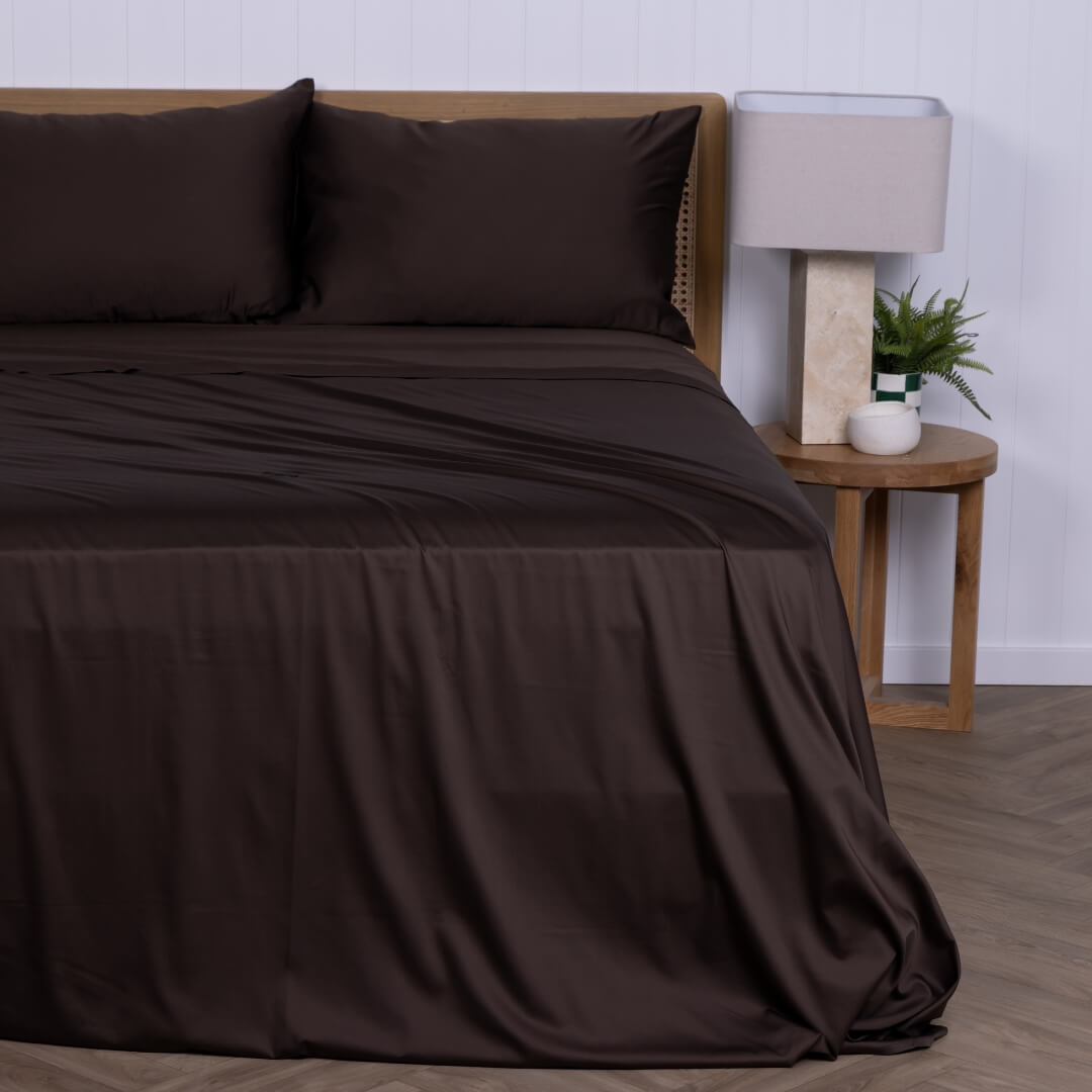 Chocolate brown bed sheets on a bed with a wooden nightstand and lamp in the background.