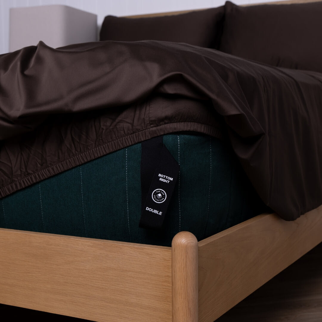 Brown fitted sheet on a wooden bed with a labelled corner loop strap.