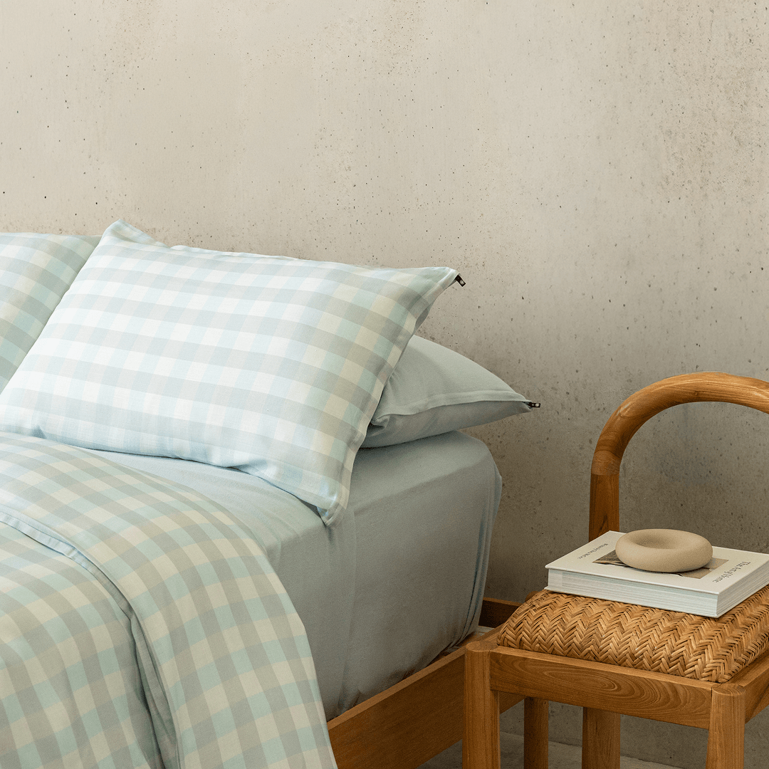 Close-up of a light blue gingham quilt cover and pillowcases layered with soft blue fitted sheets, styled beside a woven wooden chair in a modern bedroom setting