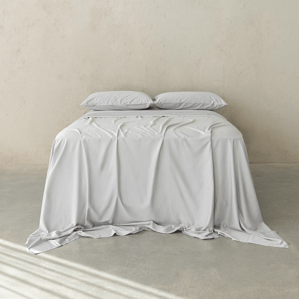 Front view of a bed dressed in light gray bedding with two matching pillows and a softly draped flat sheet against a neutral concrete wall.