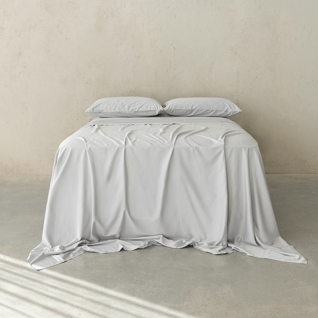 Front view of a bed dressed in light gray bedding with two matching pillows and a softly draped flat sheet against a neutral concrete wall.