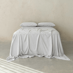 Front view of a bed dressed in light gray bedding with two matching pillows and a softly draped flat sheet against a neutral concrete wall.