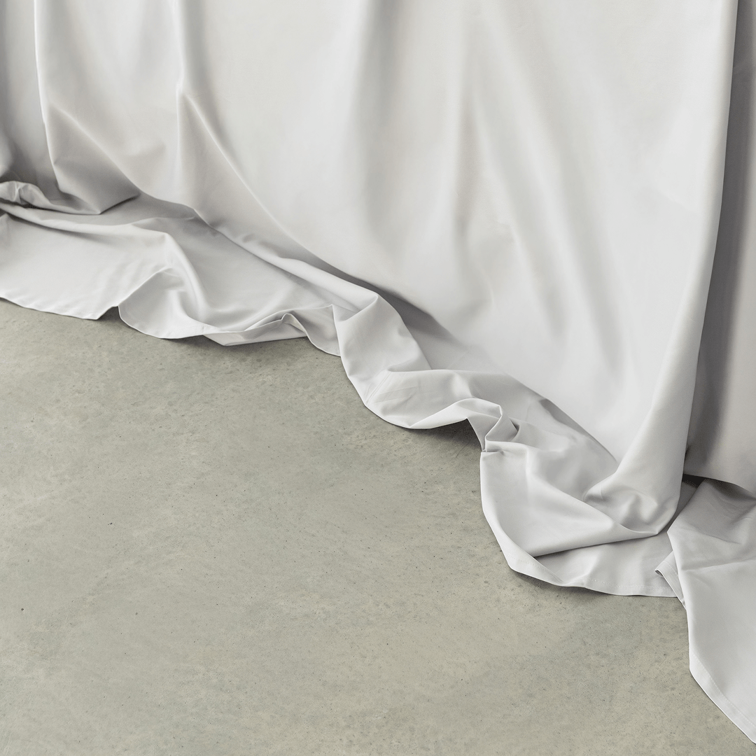 Close-up of a light gray flat sheet draping softly onto a concrete floor, showing its smooth texture and flowing folds.