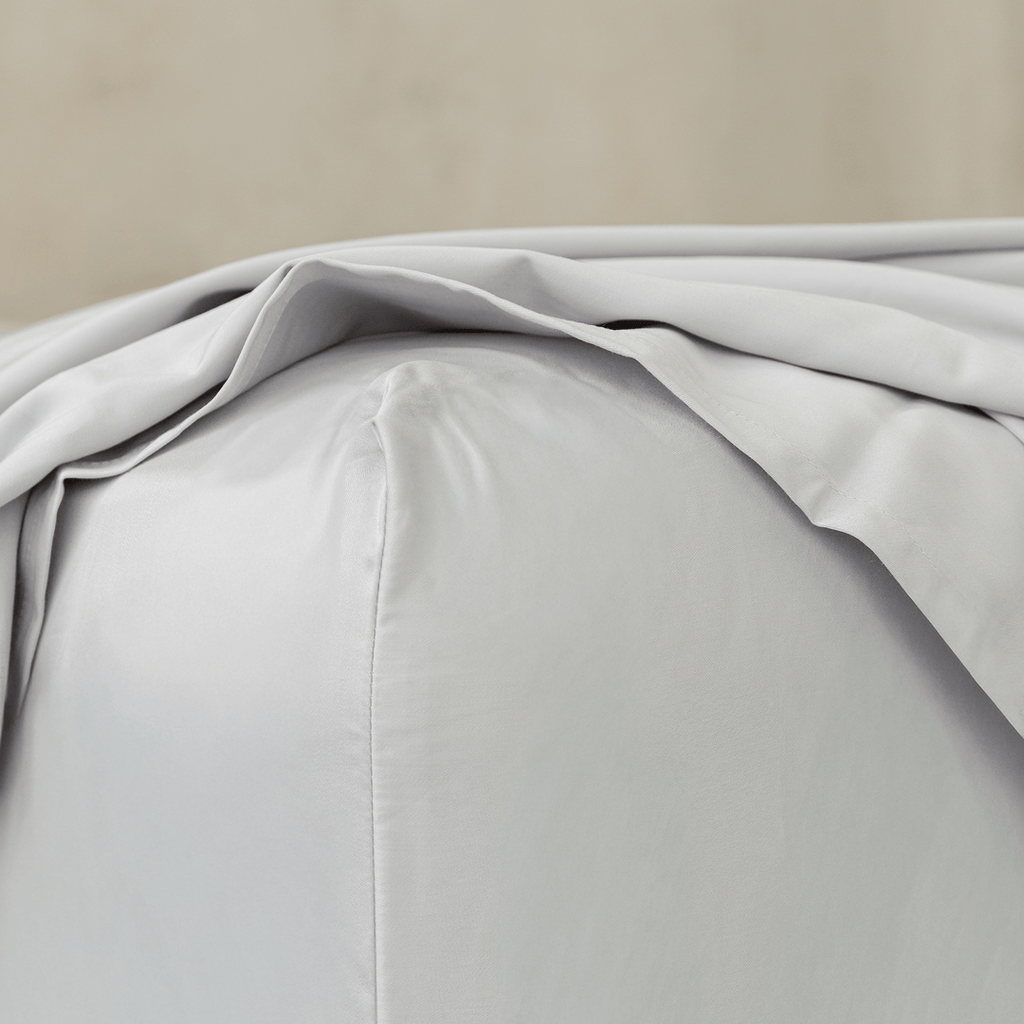 Close-up of a light gray fitted sheet stretched over a mattress corner, highlighting the smooth fabric, stitched seam, and snug fit.
