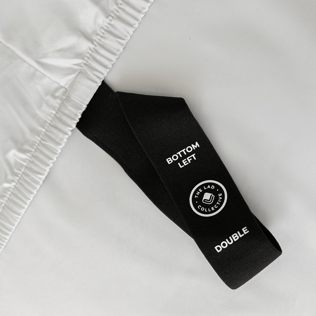 Close-up of a black fitted-sheet corner strap labelled “Bottom Left” and “Double” against light gray bedding.
