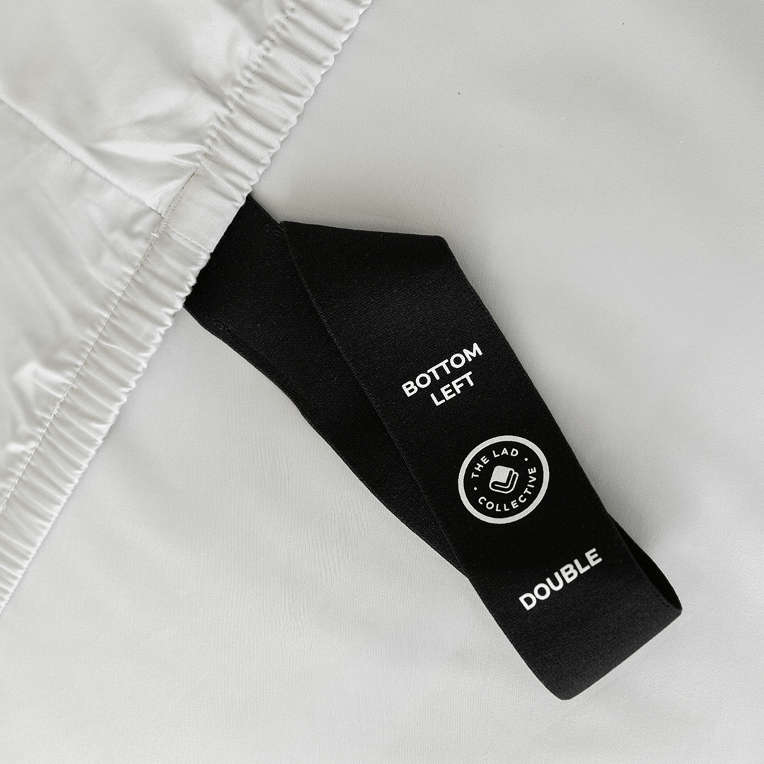 Close-up of a black fitted-sheet corner strap labelled “Bottom Left” and “Double” against light gray bedding.