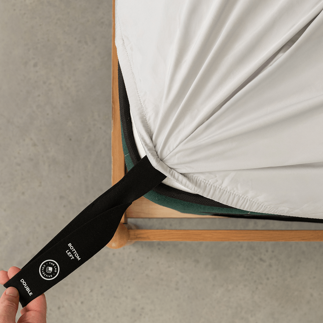 A hand pulls the labelled black corner strap of a light gray fitted sheet around the bottom-left corner of a double mattress.