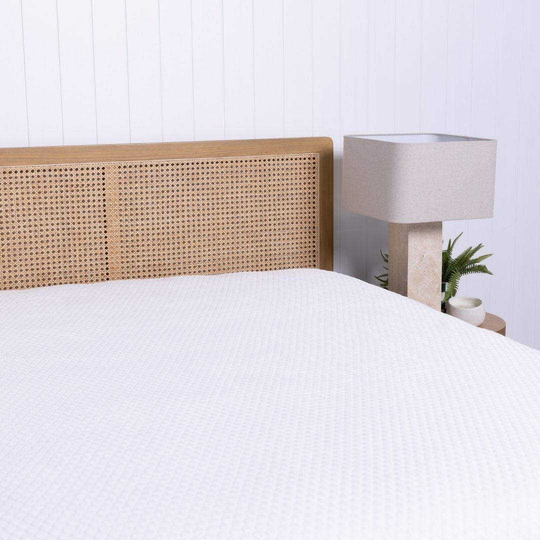 Bed with wicker headboard and white mattress protector, nightstand with lamp in the corner.