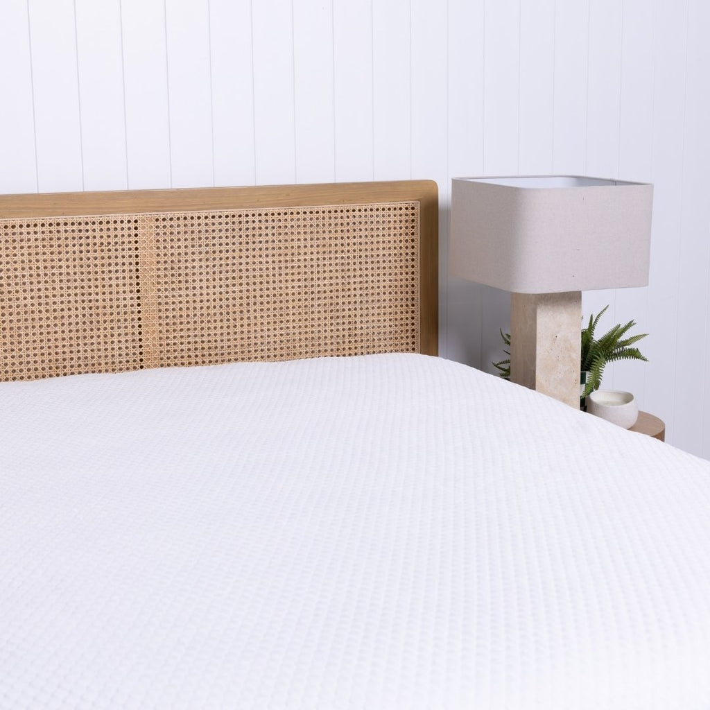 Bed with wicker headboard and white mattress protector, nightstand with lamp in the corner.