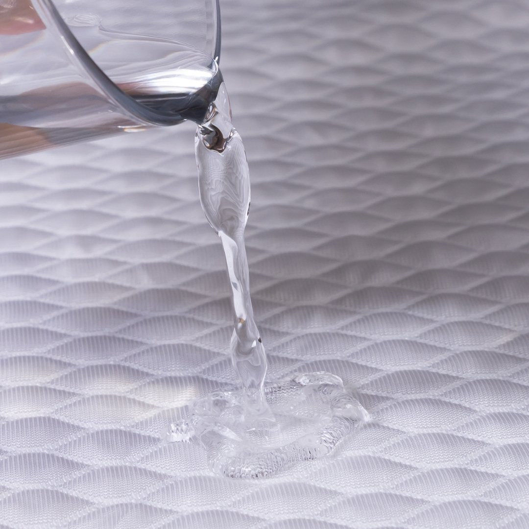 Clear liquid being poured from a glass container onto a mattress protector, showing the water resistant nature.