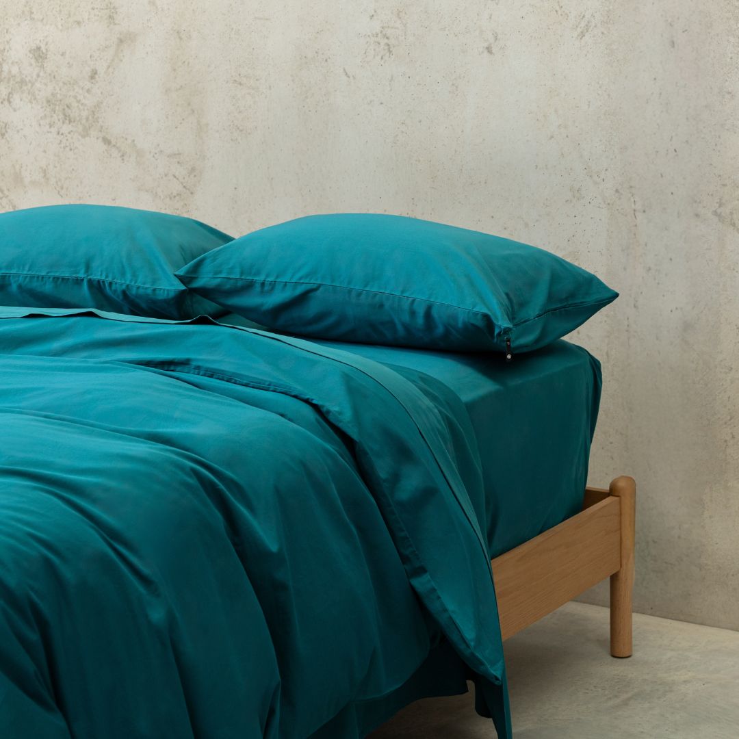 Teal bed sheets and pillows on a wooden bed against a neutral wall.