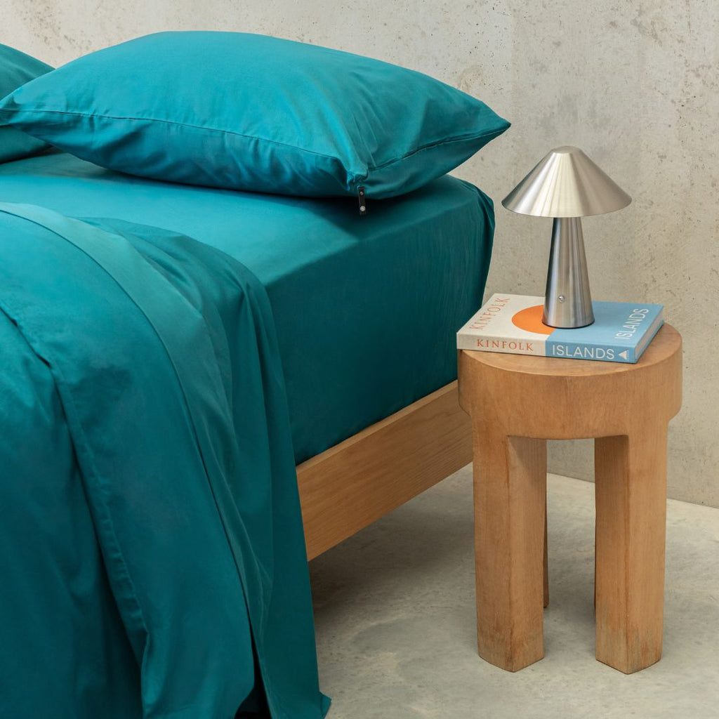 Teal bedspread on a bed with a wooden side table and lamp.