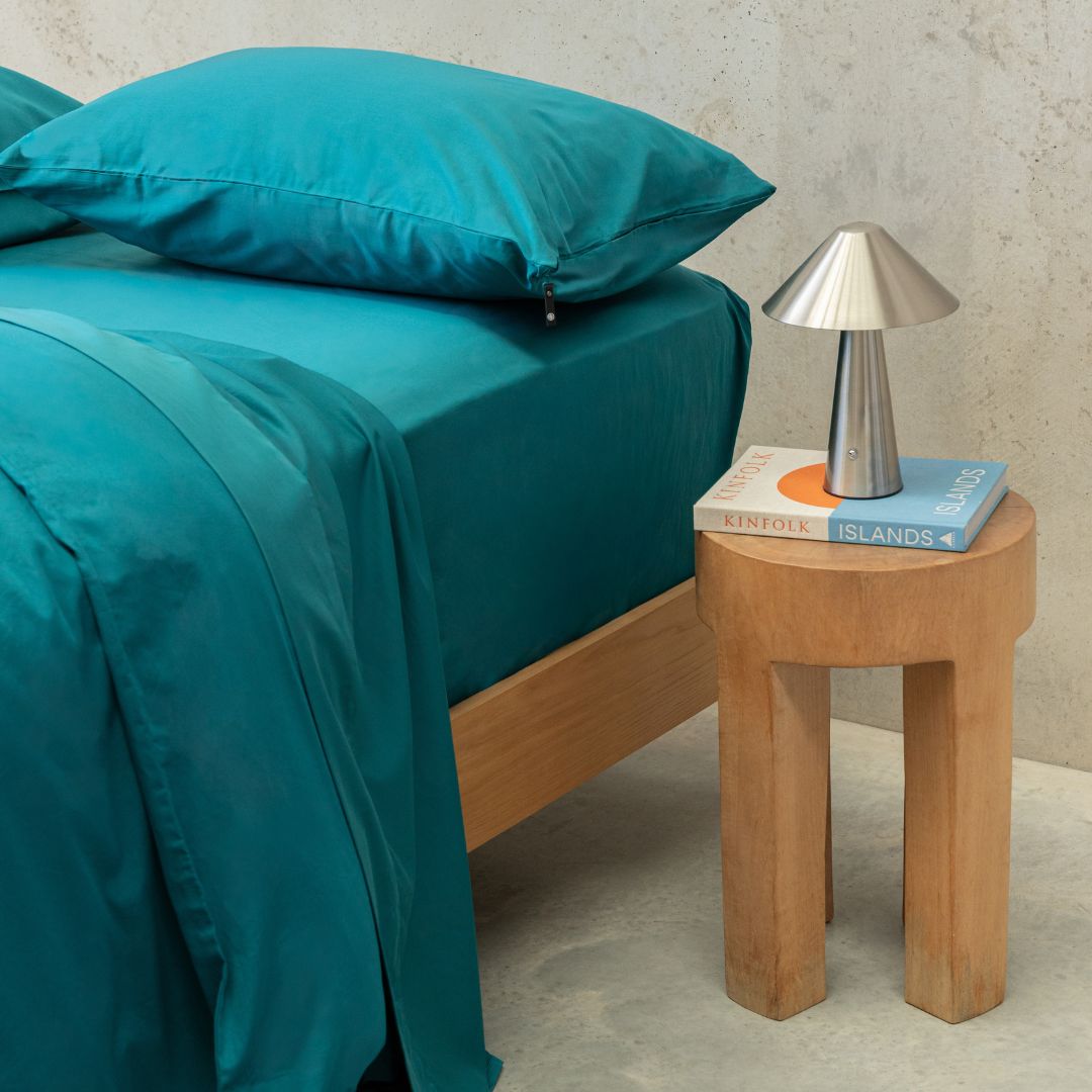 Teal bedspread on a bed with a wooden side table and lamp.
