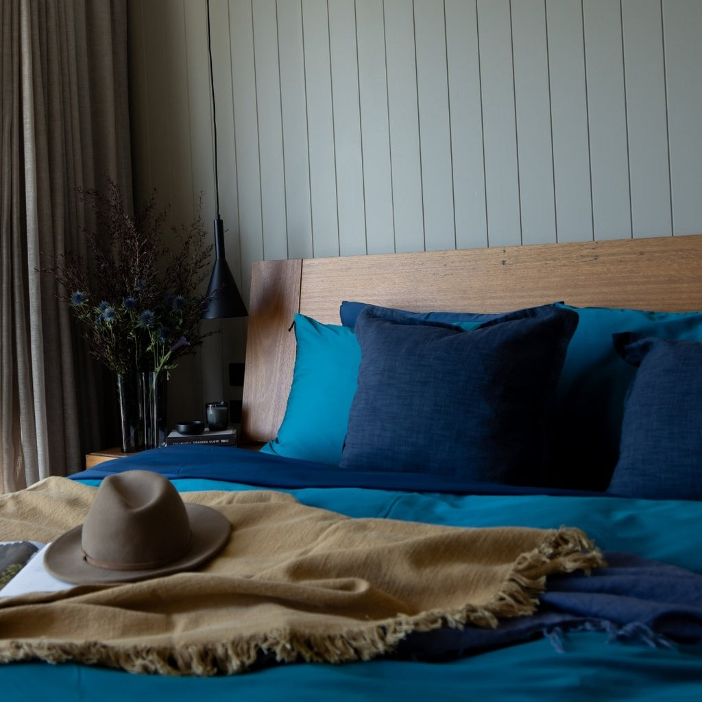 Bed with blue & teal bedding and pillows, beige blanket, and a hat on a wooden headboard.