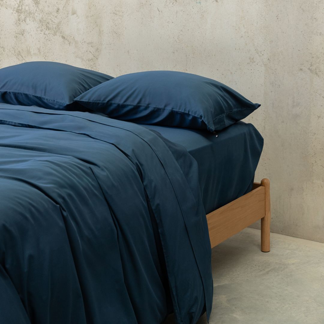 Bed with dark blue bedding and wooden headboard against a neutral wall.