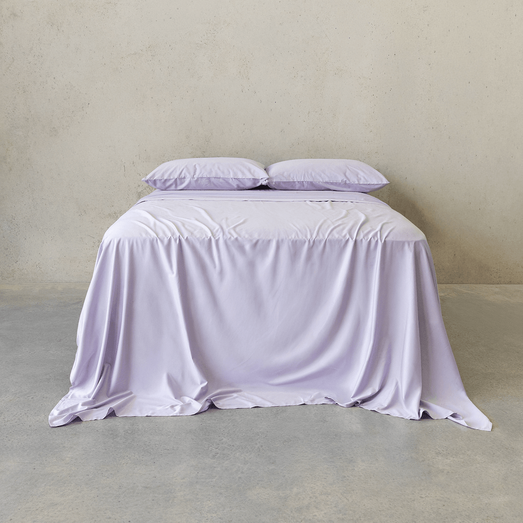 Front view of a bed dressed in a light lavender sheet set with two matching pillows, styled against a neutral concrete wall.