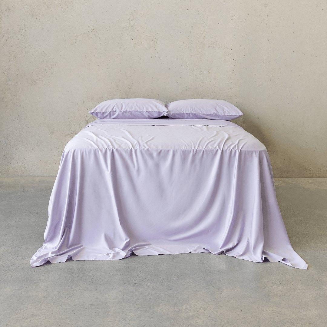 Front view of a bed dressed in a light lavender sheet set with two matching pillows, styled against a neutral concrete wall.