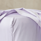 Close-up of a light lavender fitted sheet stretched over a mattress corner, showing the smooth fabric and stitched finish.