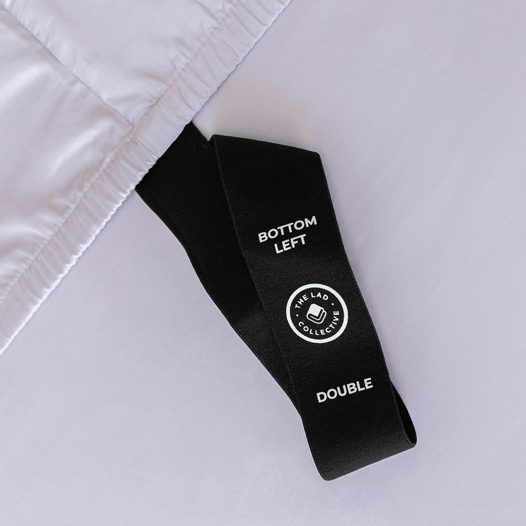 Close-up of a black fitted-sheet corner strap labelled “Bottom Left” and “Double” against light lavender fabric.