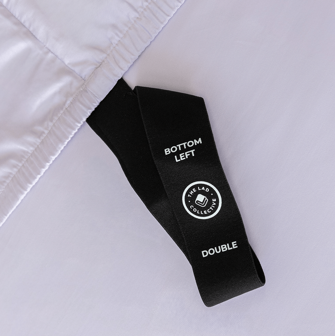 Close-up of a black fitted-sheet corner strap labelled “Bottom Left” and “Double” against light lavender fabric.
