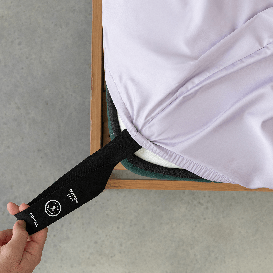 A hand pulls the labelled black corner strap of a light lavender fitted sheet around the bottom-left corner of a double mattress.
