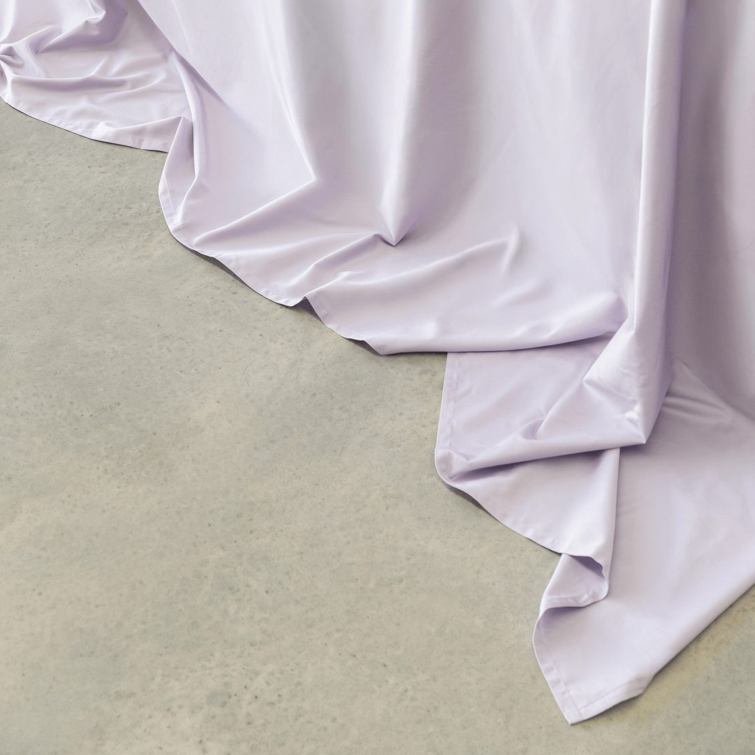 Close-up of a light lavender flat sheet draping softly onto a concrete floor, showing its smooth finish and flowing folds.