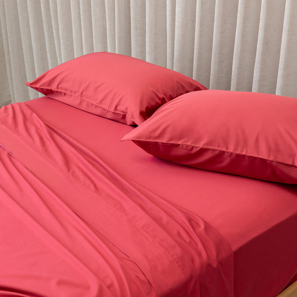 Red bed sheets and pillows on a bed with light gray curtains in the background.
