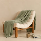 Sage green chair with a green blanket draped over it, accompanied by a mug and newspaper on a beige surface.