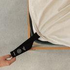 Beige fitted sheet with an extended orientation tag at the mattress corner, demonstrating the easy-to-locate labeling system for faster bed setup.