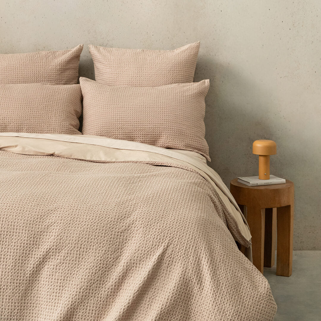 Beige bedding set with pillows on a bed against a neutral wall with a wooden side table.