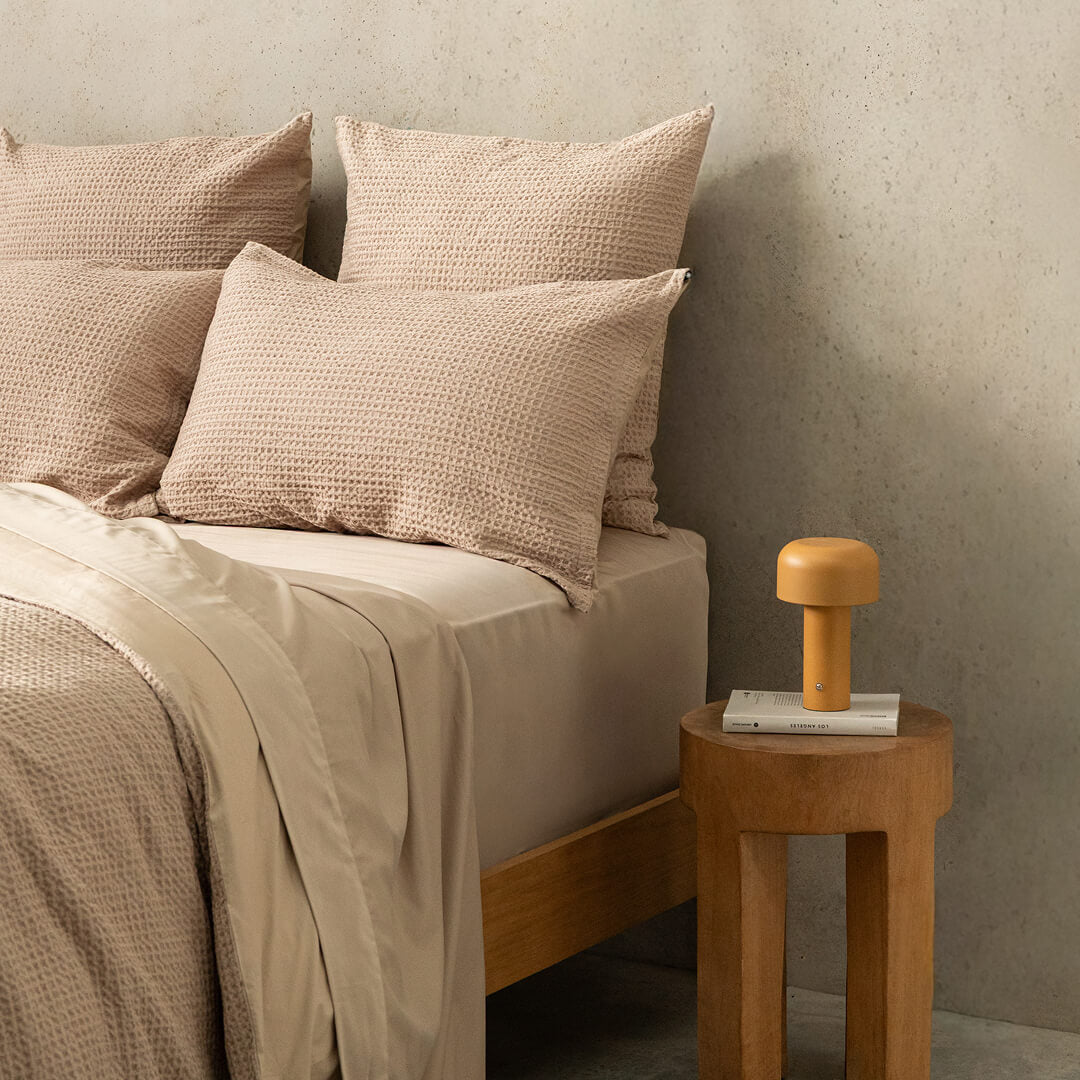 Beige bedding with pillows on a bed next to a wooden side table with a lamp.