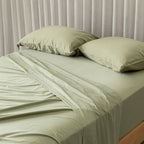 Green bed sheets and pillows on a bed with a neutral curtain background.
