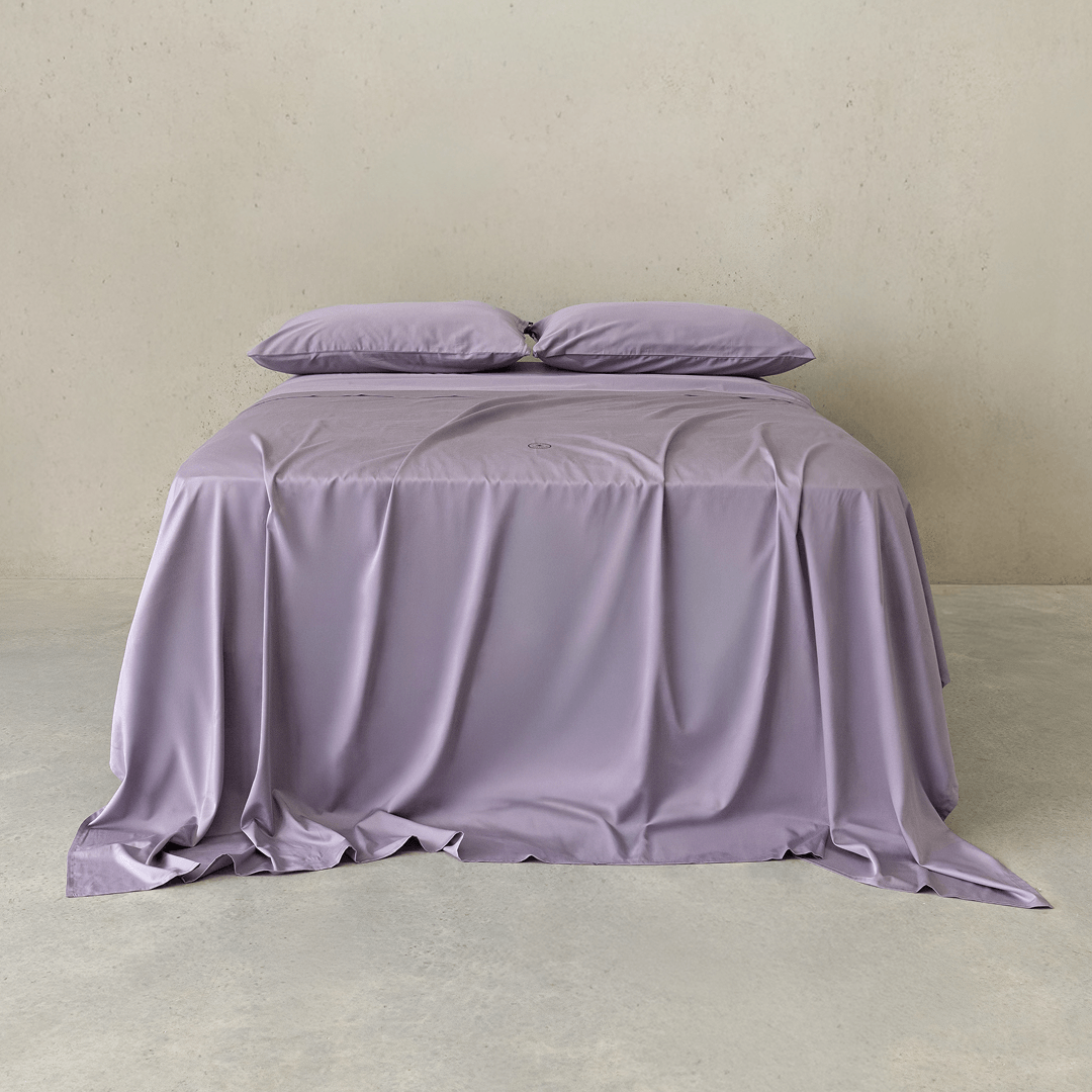 Lavender Daylesford bedding set draped over a bed with matching pillowcases, showcasing the silky fabric and relaxed, elegant styling.