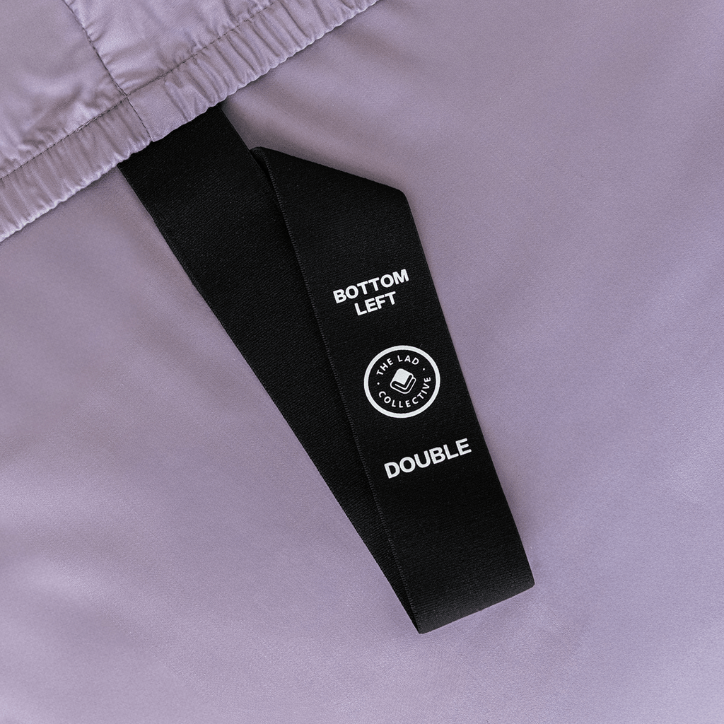 Close-up of a black corner strap attached to a lavender fitted sheet, labelled “Bottom Left” and “Double” for easy bed placement.