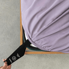 A hand pulls the labelled black corner strap of a lavender fitted sheet around the bottom-left corner of a double mattress.