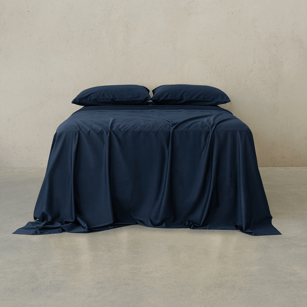 Navy blue bed sheet set draped over a double bed with two matching pillowcases against a textured wall