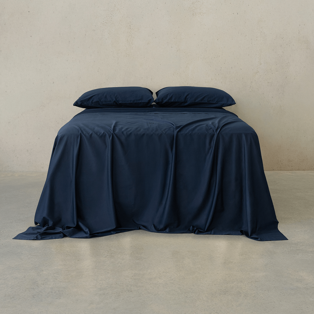 Navy blue bed sheet set draped over a double bed with two matching pillowcases against a textured wall