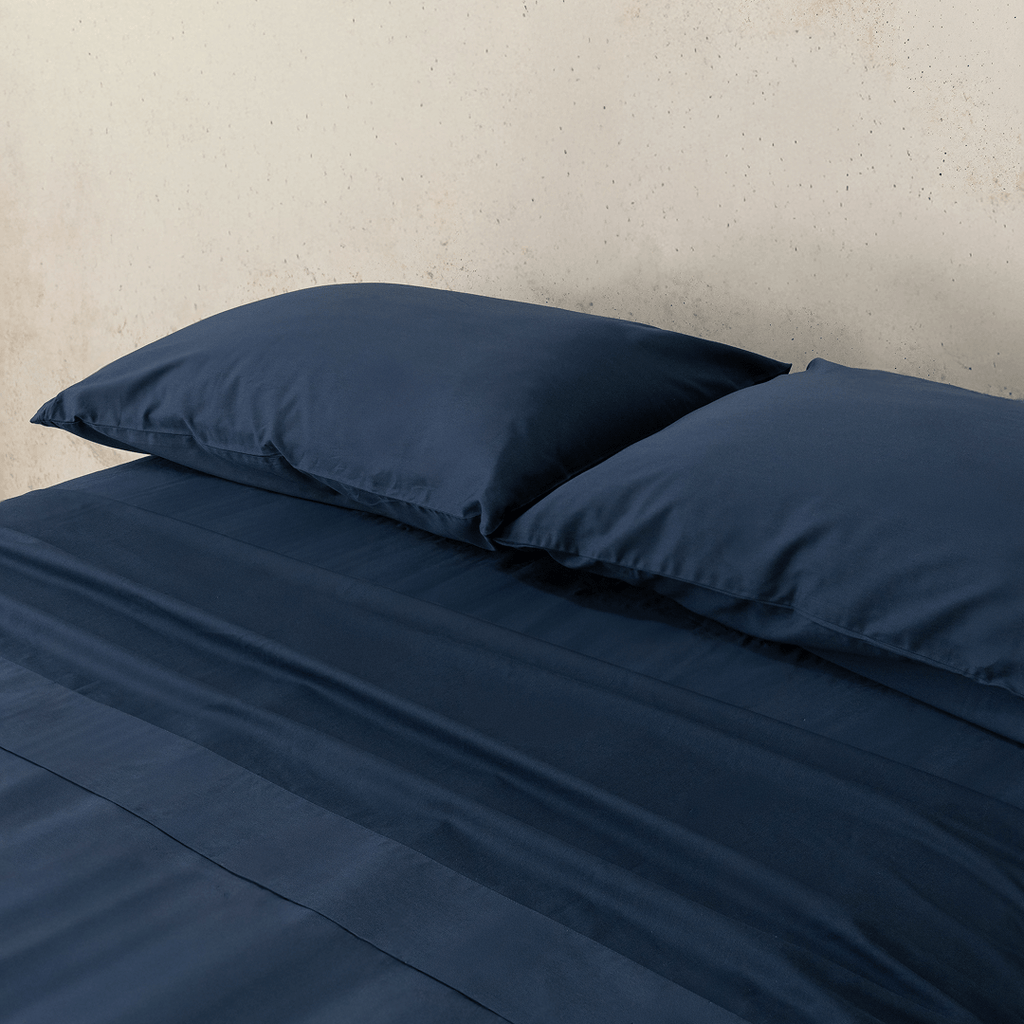 Angle view of two navy blue pillowcases and a matching flat sheet neatly arranged on a bed
