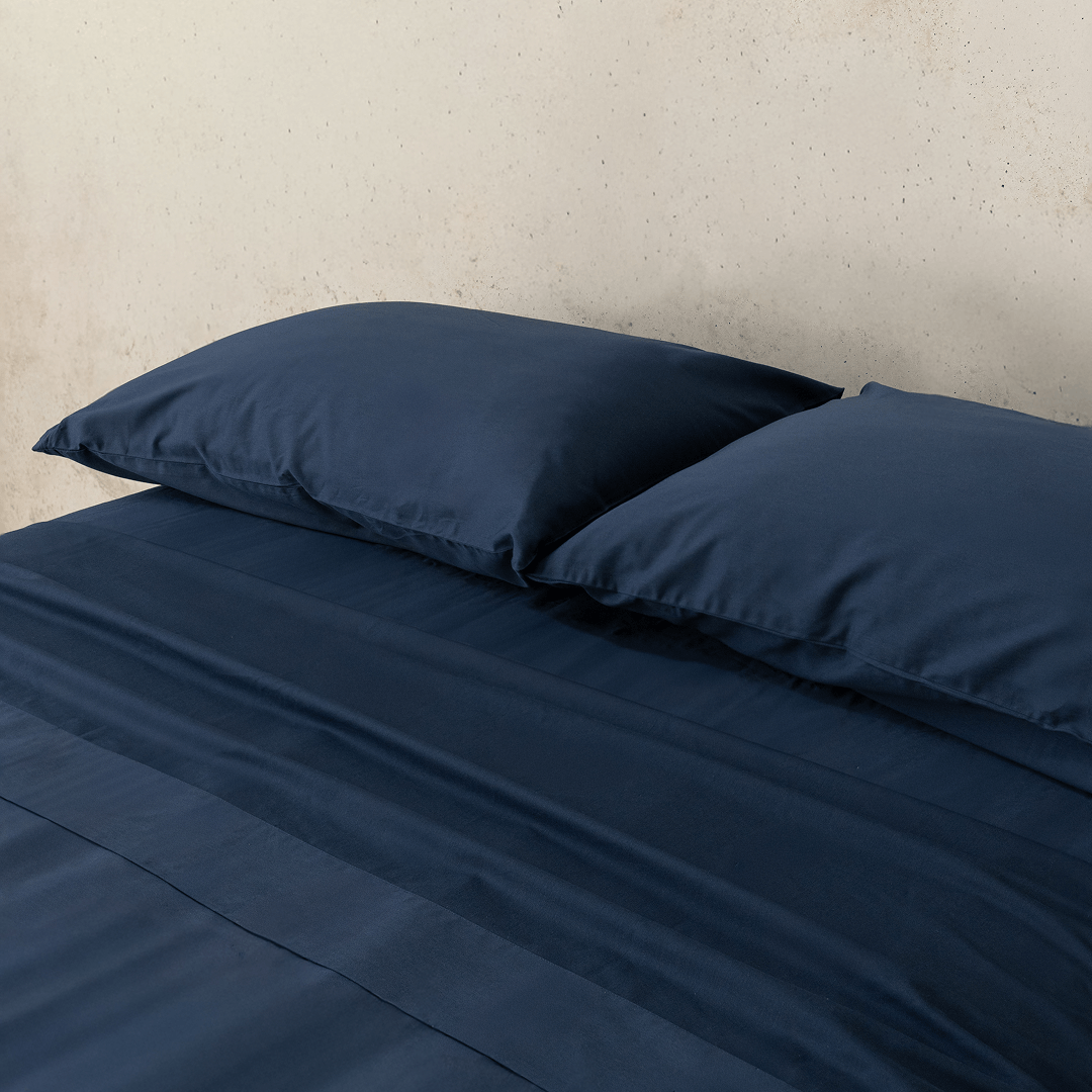 Angle view of two navy blue pillowcases and a matching flat sheet neatly arranged on a bed