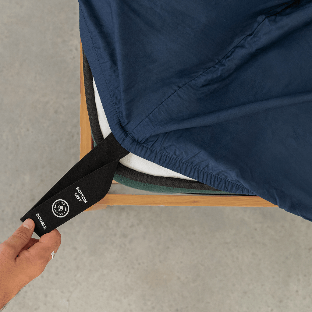 Hand pulling a black elastic corner band labeled 'Bottom Left' while securing a navy blue fitted sheet onto a mattress corner