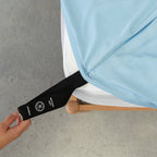 Pastel blue fitted sheet labelled corner loop strap being pulled onto bed.