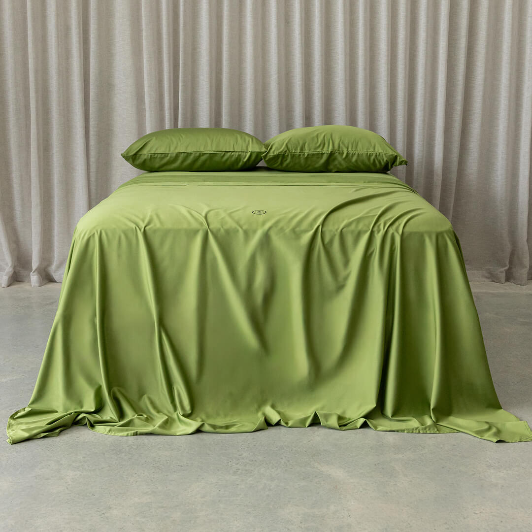 Green bedspread on a bed with matching pillows against a neutral curtain background.