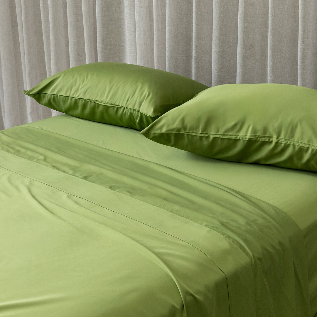 Green bed sheets and pillows on a bed with a neutral curtain background.